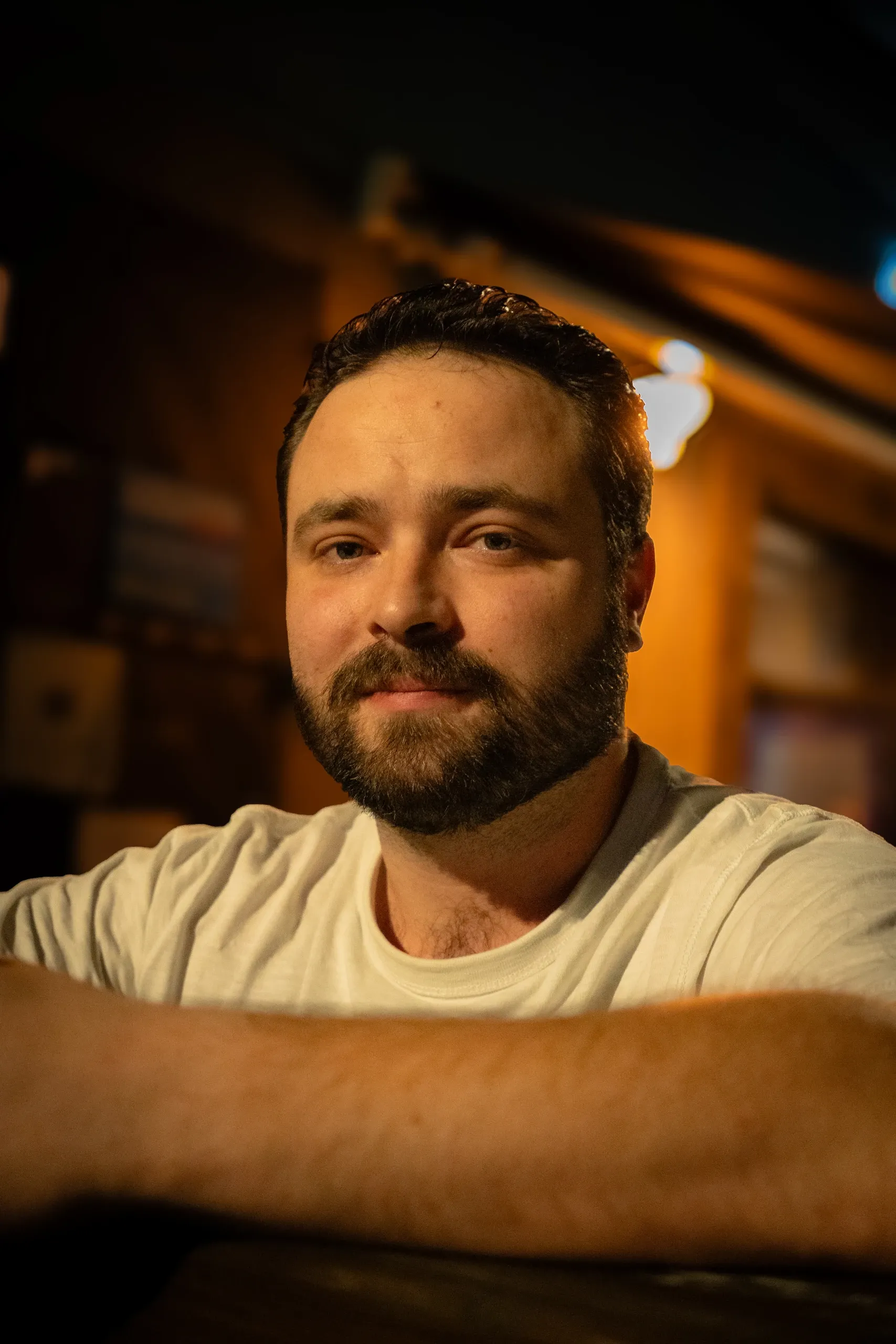 Portrait d'un homme barbu en t-shirt blanc, accoudé au comptoir d'un bar à la lumière chaude.