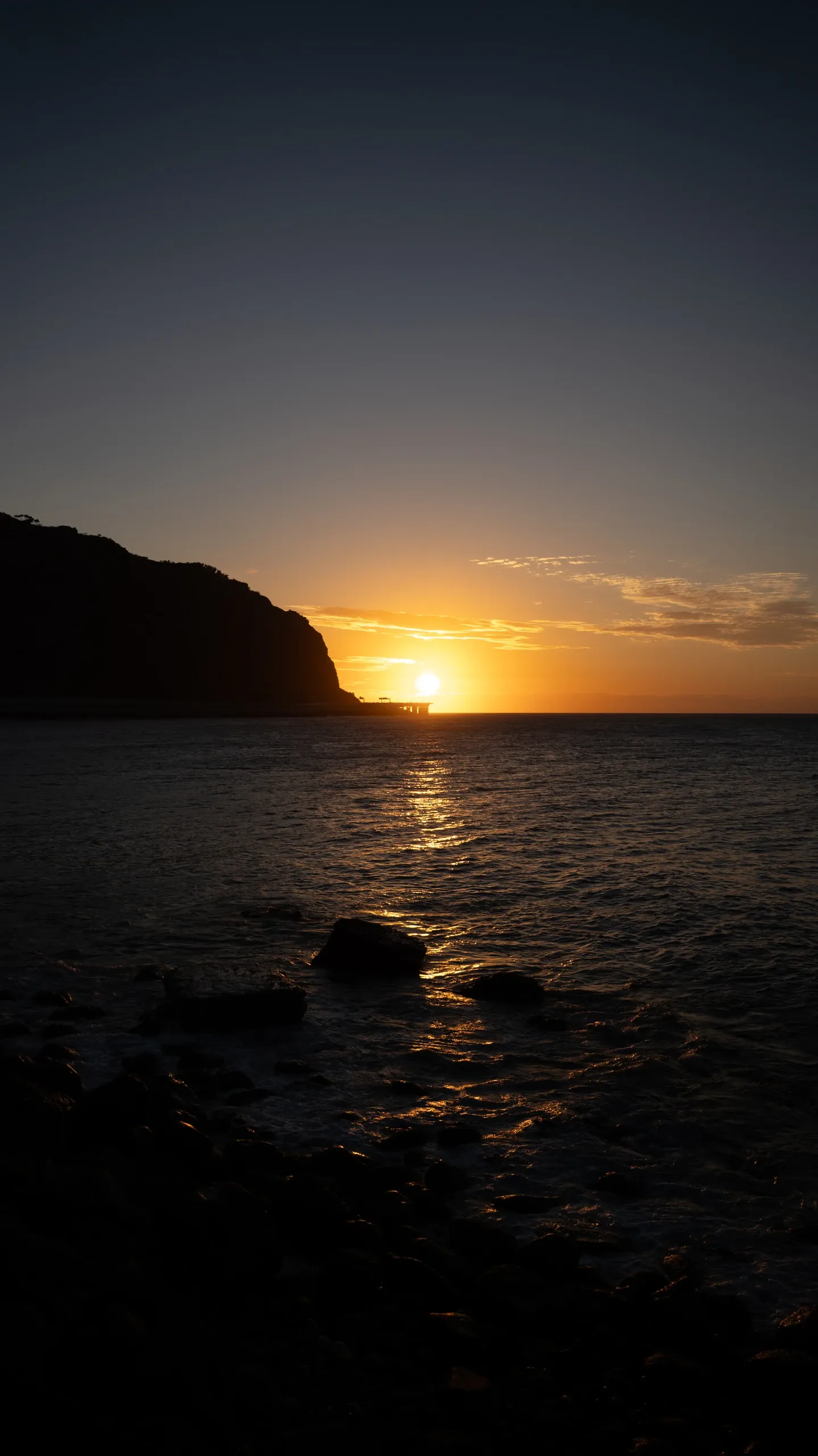 Le soleil touche l'horizon, entre une falaise sombre et une mer bleu nuit.