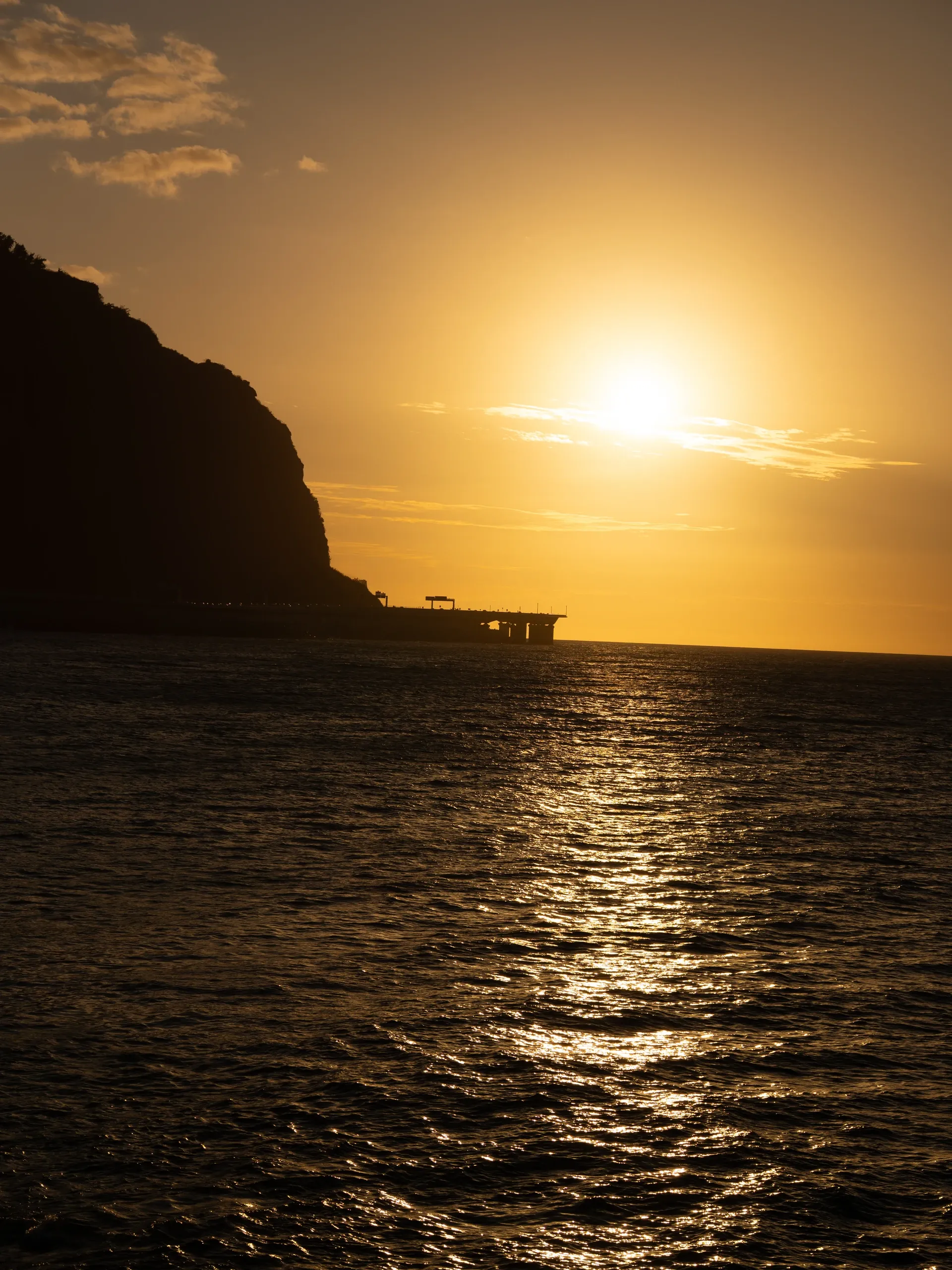 Soleil doré au-dessus de l'océan au Barachois, une falaise en silhouette à gauche.