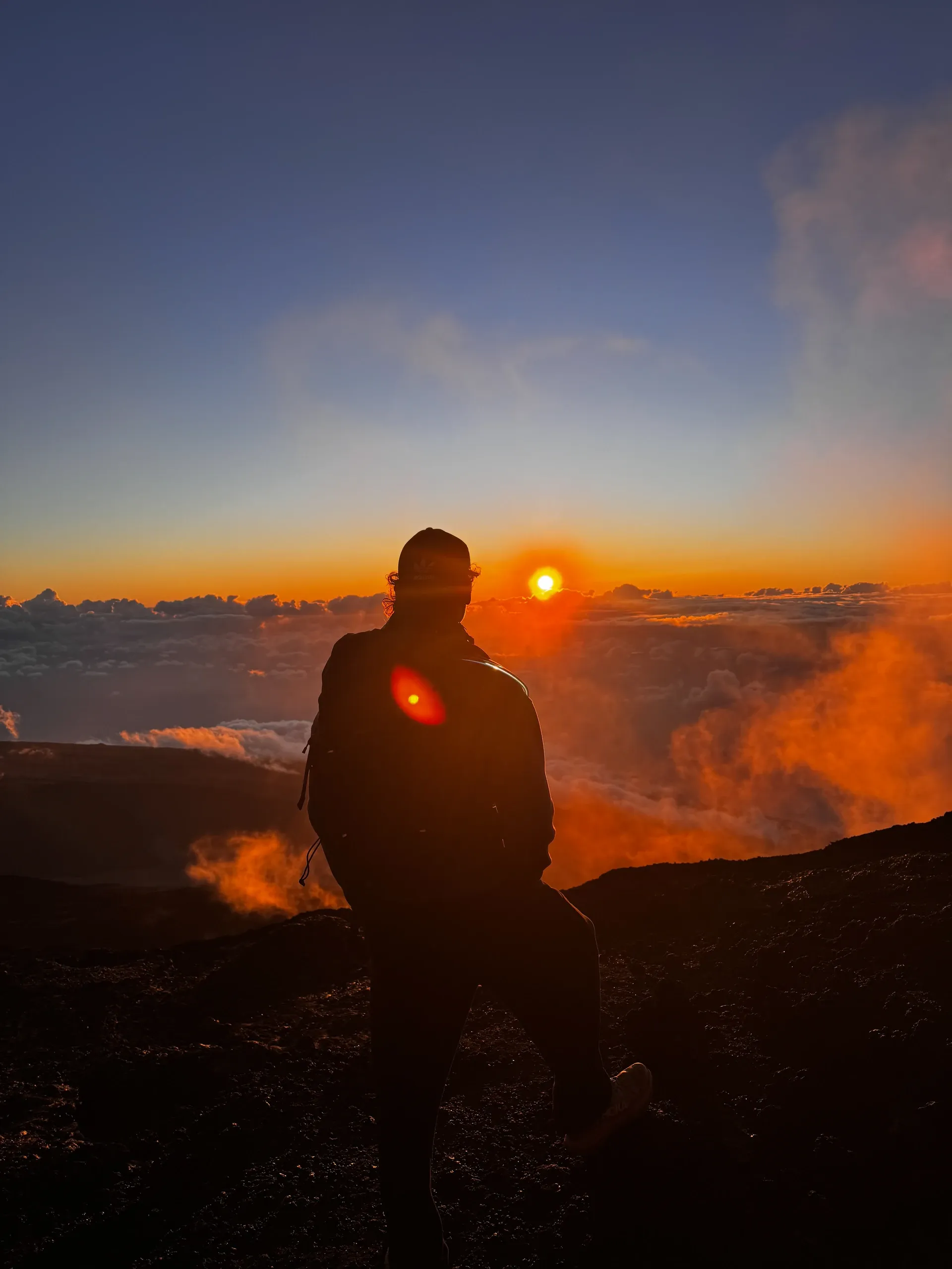 Silhouette de Dono face au soleil levant, au-dessus d'une mer de nuages.