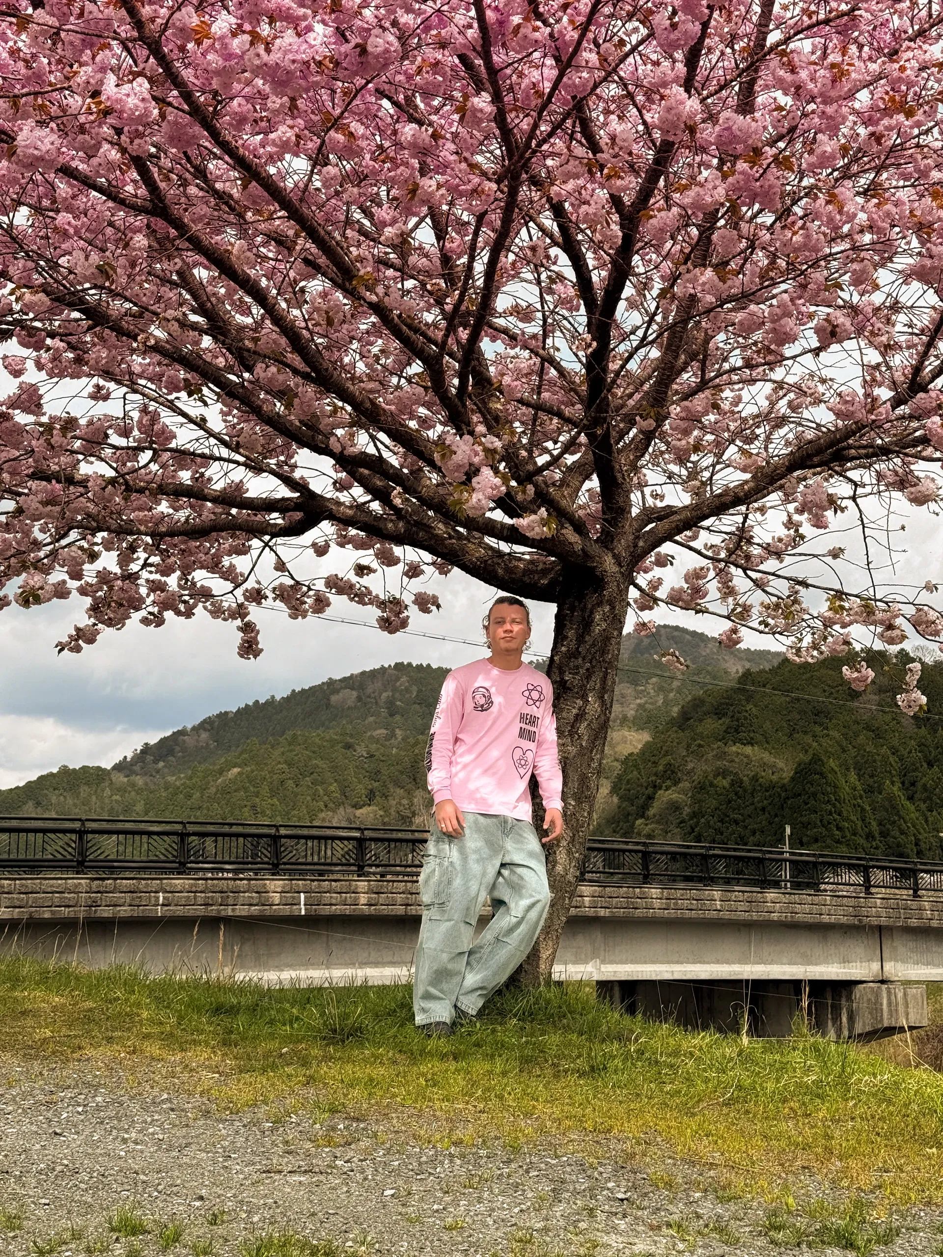 Dono en sweat rose, debout sous un cerisier en fleurs au bord d'une rivière.
