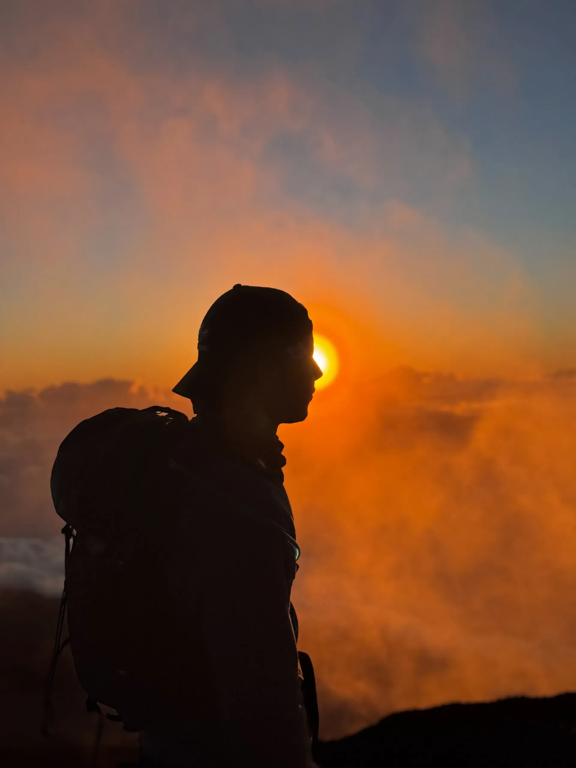 Profil de Dono en casquette et sac à dos, à contre-jour dans les nuages orangés.